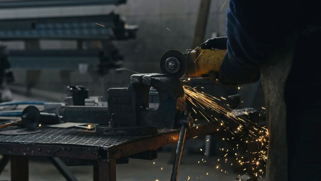 A worker uses an angle grinder on a metal piece, creating a shower of bright sparks.